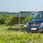 Preview: Outwell Hillcrest Tarp am Mercedes-Van, grün gespannt, zwei Personen sitzen darunter auf Campingstühlen im Gras