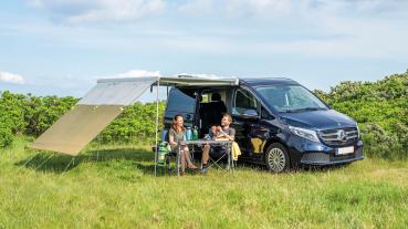 Outwell Hillcrest Tarp am Mercedes-Van, grün gespannt, zwei Personen sitzen darunter auf Campingstühlen im Gras