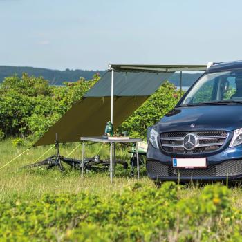 Outwell Hillcrest Tarp am Mercedes-Van, grün gespannt, zwei Personen sitzen darunter auf Campingstühlen im Gras