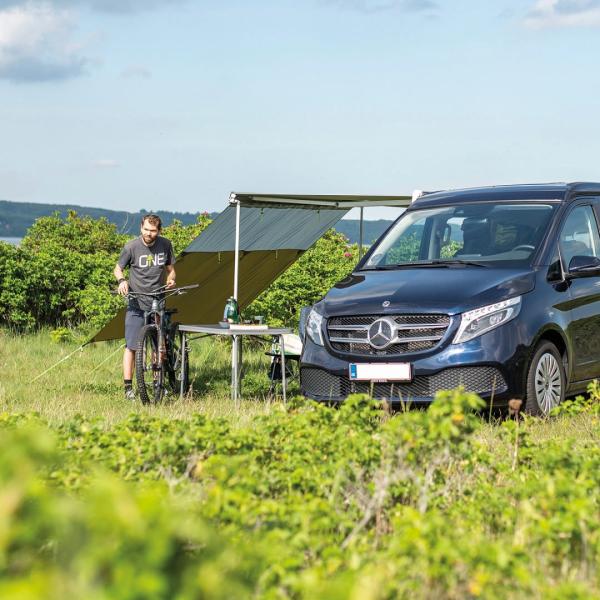 Outwell Hillcrest Tarp am Mercedes-Van, grün gespannt, zwei Personen sitzen darunter auf Campingstühlen im Gras